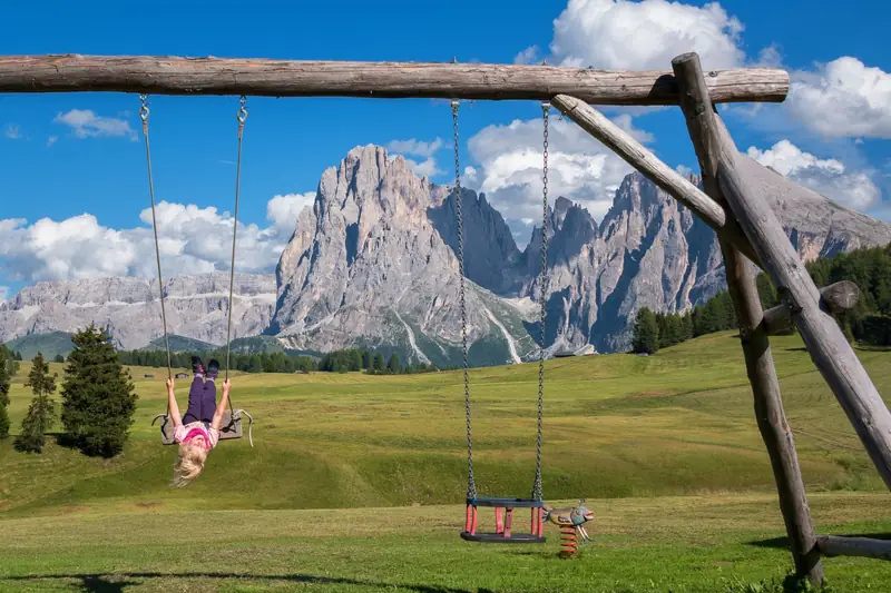 Ein Kind auf einer Holzschaukel auf einer Almwiese vor den Dolomiten
